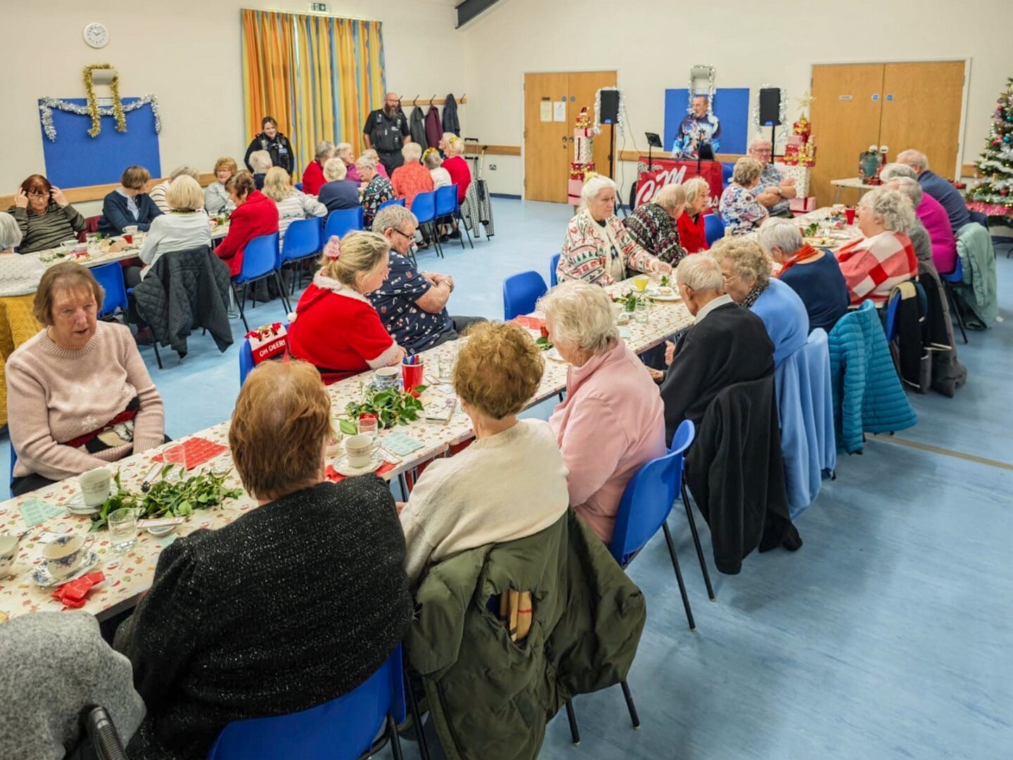 A wide view of a well-attended community tea party inside Millennium Hall, showing residents seated at long decorated tables with refreshments.
