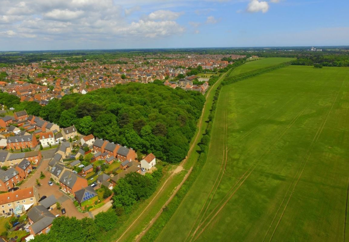 Aerial view of Kesgrave showing houses, woodland, and green open fields on the town’s edge under a blue sky.