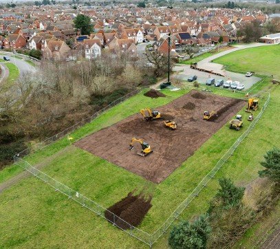 Aerial view of Kesgrave Pump Track construction. Tru7 Group excavators clear a rectangular earth patch at Millennium Sports Ground, surrounded by security fencing with a residential neighbourhood and green field in the background.