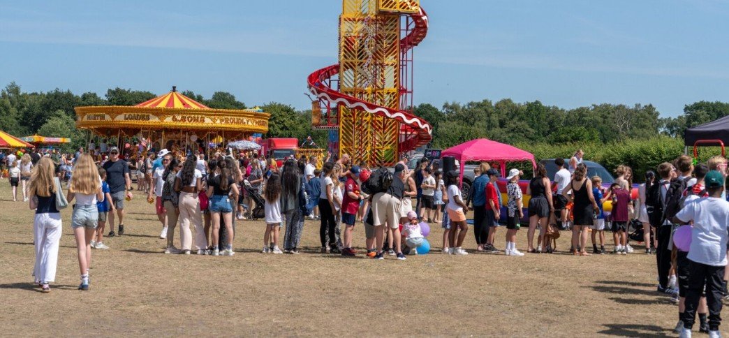 A wide, sunny shot of the Kesgrave Fun Day at Millennium Sports Field, showing families enjoying fairground rides like a golden carousel and a tall spiral slide under a clear blue sky.